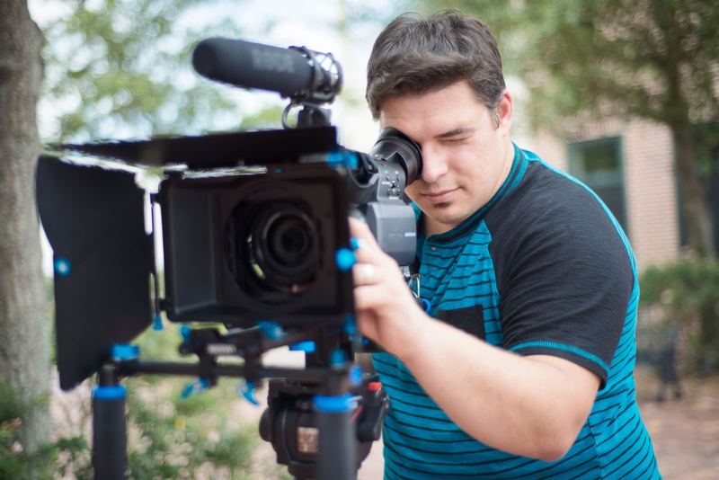 Young male videographer using a professional video camera outdoors with a blurred background.